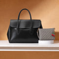 A black leather handbag with double top handles sits on a white shelf against a warm brown background, next to a gray quilted wristlet pouch with a red cherry charm. Bag by HBH