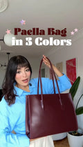 A woman in a light blue shirt holds up a large leather tote bag in a deep burgundy color. The image has text overlays reading 'Paella Bag in 3 Colors' with pink stars, set against an indoor home background.